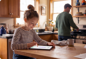 A child using a tablet at a kitchen counter while an adult prepares food in the background